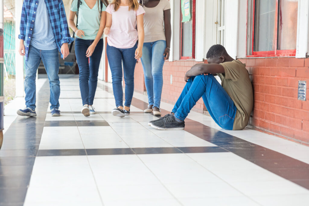 male teenager upset seated in school hallway while group of teenagers walk towards him